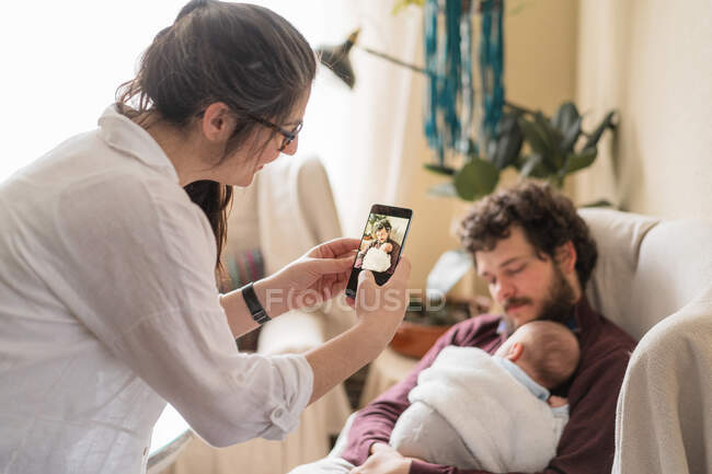 Зміст жінки, яка фотографує коханого чоловіка з невпізнаваною новонародженою дитиною на мобільному телефоні в кімнаті — стокове фото