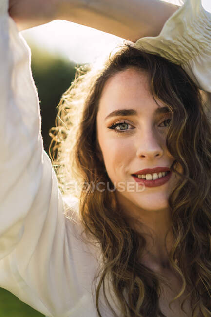 Beautiful content female with blue eyes and long curly hair raising arms gracefully and looking at camera with smile while spending sunny day in verdant nature — Stock Photo
