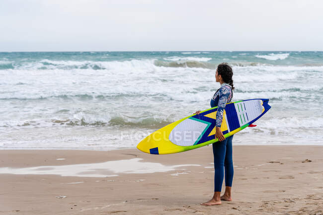 Athlète féminine ethnique joyeuse en combinaison avec kiteboard debout sur le rivage sablonneux contre cerf-volant gonflable — Photo de stock
