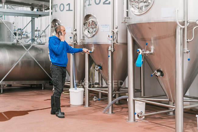 Вид збоку чоловічий підприємець говорить на мобільний телефон проти посудин з нержавіючої сталі на мокрій підлозі на заводі пива — стокове фото