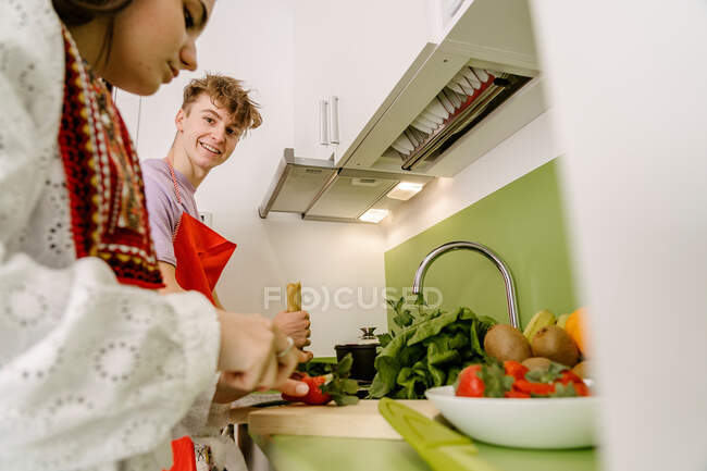 Low angle of crop ethnic female in stylish outfit cutting fresh strawberry on chopping board while cooking in kitchen with cheerful boyfriend — Photo de stock