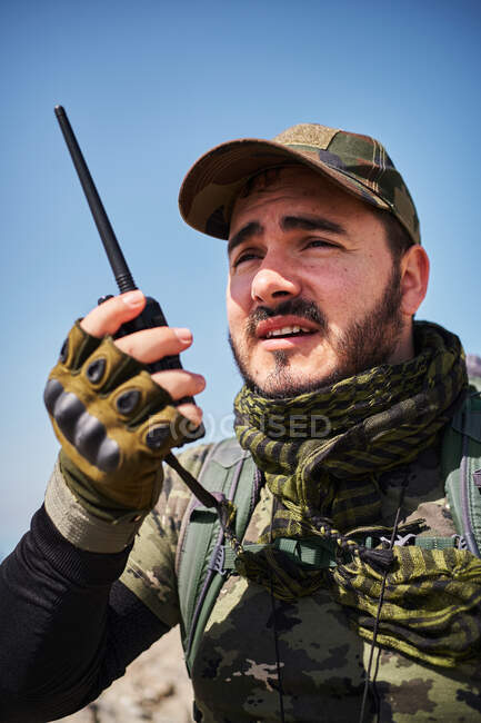 Desde abajo soldado barbudo en uniforme hablando en walkie talkie mientras mira hacia otro lado en el día soleado - foto de stock