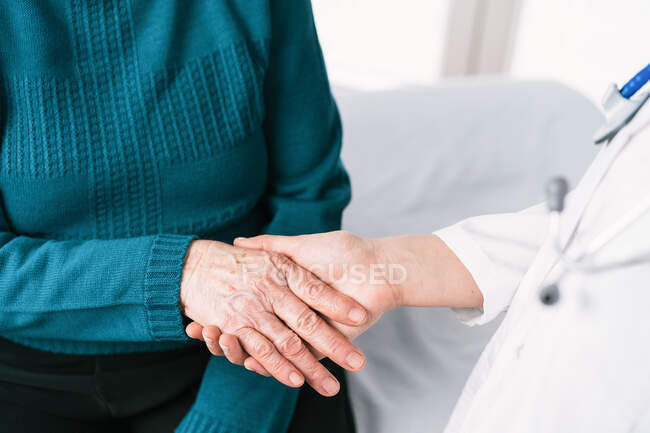 Crop médico anónimo hablando con una mujer mayor mientras se toma de la mano durante el examen en el hospital - foto de stock