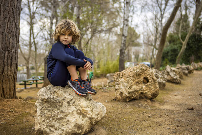 Charming child in sports clothes sitting on rough stone while looking away against trees in daytime — Foto stock