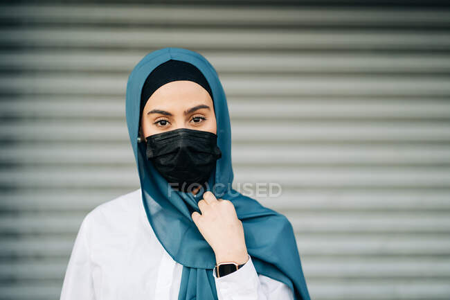 Mujer musulmana con máscara protectora y pañuelo tradicional de pie contra la pared en la ciudad y mirando a la cámara - foto de stock