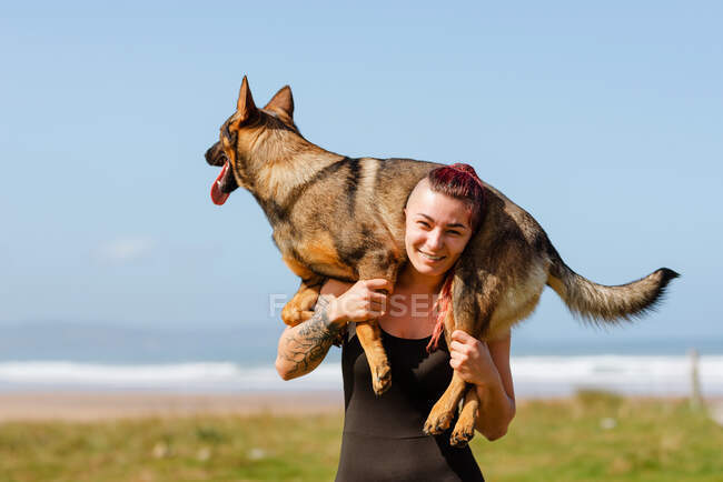 Tattooed female athlete with cute purebred dog on shoulders looking at camera on sunny day — Stock Photo