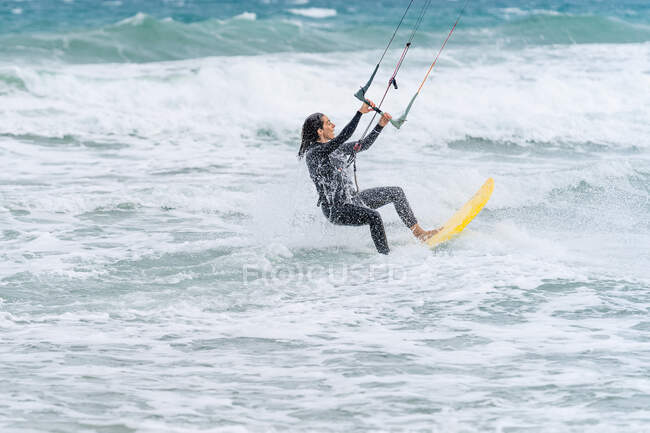 Active female athlete on kiteboard holding control bar while practicing kitesurfing and looking away on foamy ocean — Stock Photo