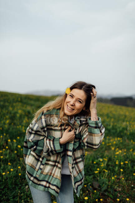 Zufriedene junge Frau im karierten Hemd blickt in die Kamera auf einer Wiese mit blühenden Blumen unter bewölktem Himmel — Stockfoto