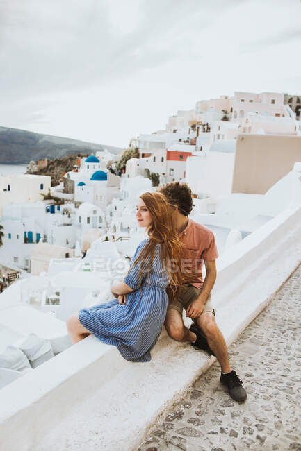 Молодая романтическая пара нежно сближается, стоя на прибрежной улице с белыми типичными домами возле синего волнистого моря в Санторини — стоковое фото