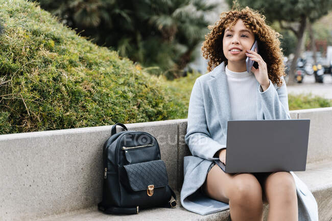 Focused young African American female in blue coat working on netbook and talking on mobile phone while sitting on stone bench in city park on clear spring day — Photo de stock