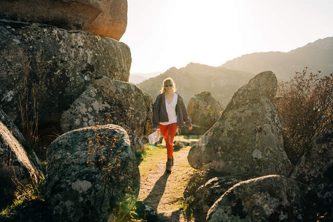 Longitud completa de contenido joven excursionista femenina con ropa elegante y gafas de sol sonriendo mientras camina en medio de enormes rocas de piedra durante el trekking en el valle montañoso - foto de stock