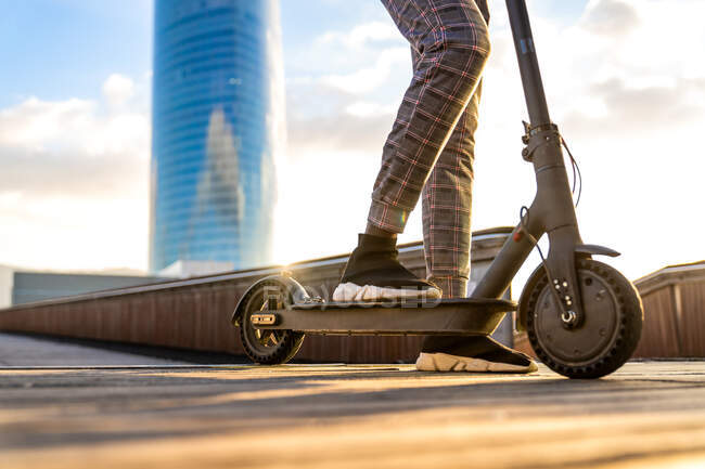 Обрізаний невпізнаваний етнічний підприємець, який їде електричним скутером на міському мостовому проході проти будівель під хмарним блакитним небом — стокове фото