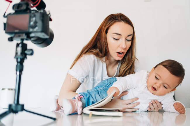 Junge Mutter umarmt und stützt kleines Baby am Tisch, während sie Video für persönlichen Videoblog im hellen Raum aufnimmt — Stockfoto
