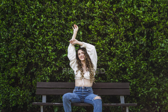 Весела приваблива жінка в повсякденному вбранні з голим животом, сидячи на дерев'яній лавці в пишному парку, піднімаючи руки витончено і дивлячись на камеру з зубною посмішкою — стокове фото