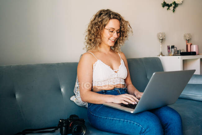 Young happy self employed woman in casual clothes and eyeglasses sitting on comfortable sofa and working remotely on laptop — Photo de stock