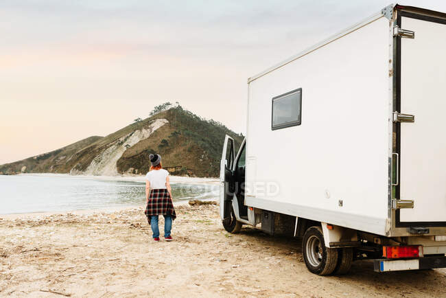 Rückansicht einer anonym reisenden Hipsterin, die in der Nähe eines geparkten Lastwagens am Ufer des Sees in bergiger Gegend steht — Stockfoto