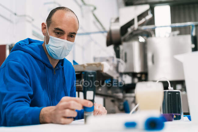 Hombre especialista en control de calidad en máscara estéril estudiando líquido alcohólico en la mesa contra equipos profesionales en fábrica - foto de stock