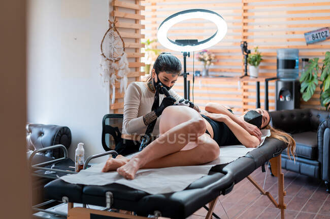 Tätowiererin in Handschuhen mit professioneller Maschine, die Tätowierung auf den Körper der Frau im Salon aufträgt — Stockfoto