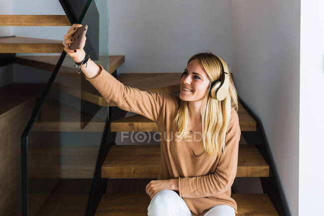 Mujer positiva escuchando música en auriculares y tomando fotos en el teléfono inteligente mientras está sentada en escaleras de madera en casa y entretenida durante el fin de semana - foto de stock