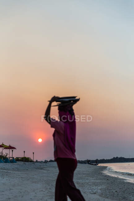 Visão traseira fêmea anônima em lenço de cabeça carregando coisas na cabeça e andando em litoral arenoso durante o pôr do sol pitoresco — Fotografia de Stock