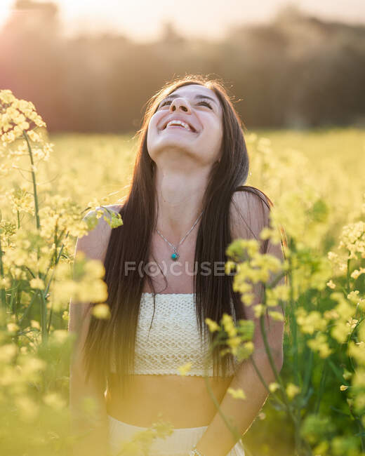 Entzückte junge Brünette in weißem Top und Jeansjacke lacht fröhlich auf blühendem Rapsfeld an sonnigem Tag — Stockfoto