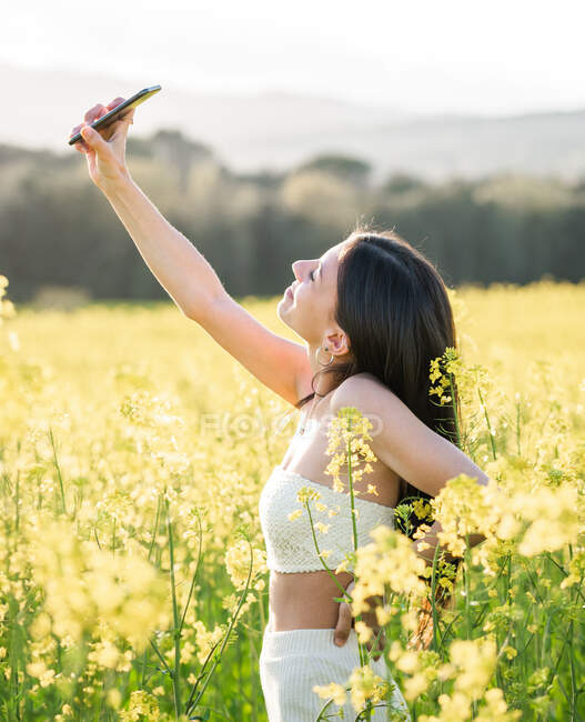 Seitenansicht lächelnde schlanke Brünette in weißer Sommerkleidung macht Selfie auf dem Smartphone, während sie bei sonnigem Wetter auf einem blühenden Rapsfeld steht — Stockfoto