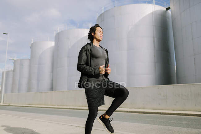 Full body ethnic man in black sportswear jogging in place while warming up on concrete street of industrial city district — Stock Photo