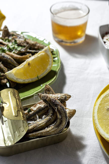 From above of delicious fried anchovies served on plate and can with lemon and placed on white table with glass of beer — Fotografia de Stock