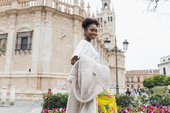 Vue de côté souriante jolie femme afro-américaine avec manteau à portée de main debout dans le quartier historique de la ville le jour du printemps chaud et regardant la caméra — Photo de stock