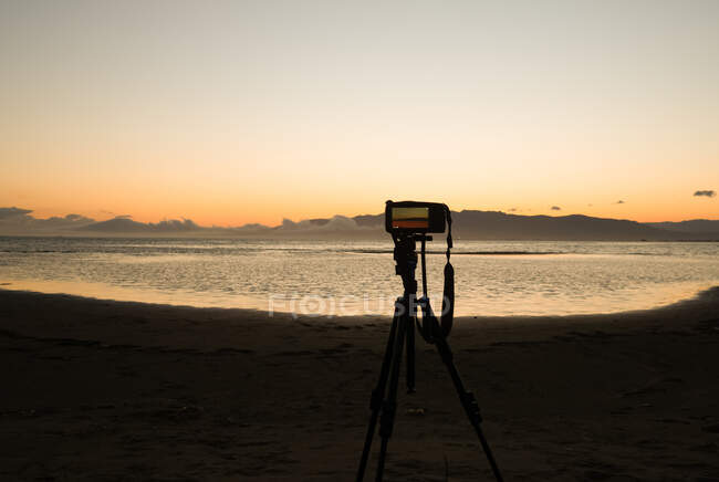 Tripod with photo camera placed on sandy beach near sea at sundown time with colorful sky — Stock Photo