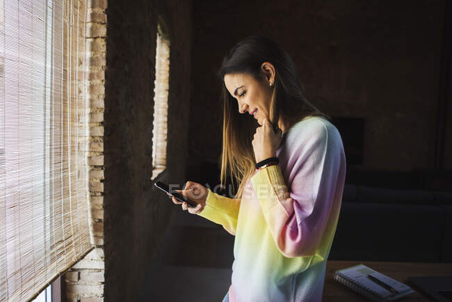 Side view of cheerful female surfing internet on cellphone while sitting on table in loft style house — Stock Photo