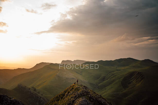 Distant tourist with dog on high peak of mountain range with slopes in green grass under clouds Txindoki, Spain — Stock Photo