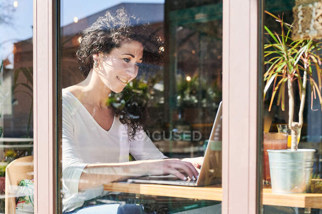 A través de la vista de cristal de una empleada remota étnica adulta trabajando en netbook en la mesa - foto de stock