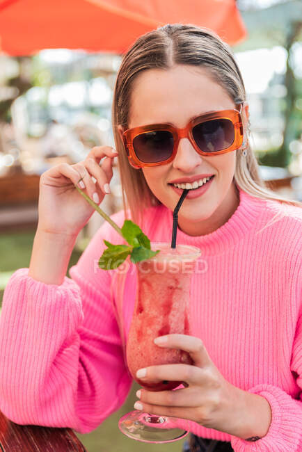 Обрізати мрійливу жінку в рожевому светрі, сидячи за столом зі смачним холодним ягідним коктейлем у відкритому барі і дивлячись на камеру — стокове фото