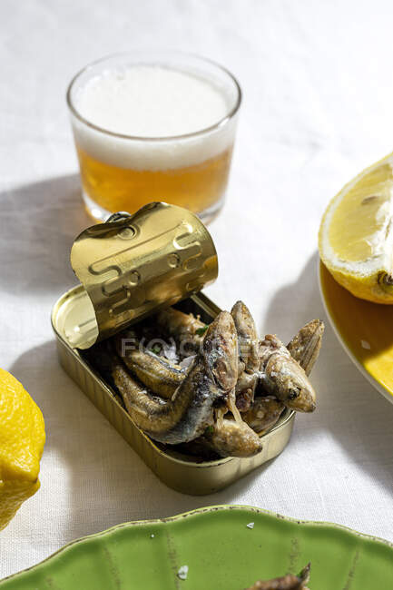 From above of delicious fried anchovies served on can with lemon and placed on white table with glass of beer - foto de stock
