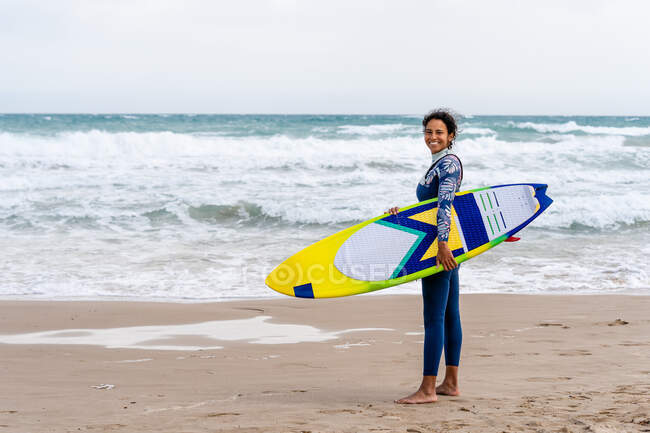 Side view of cheerful ethnic female kitesurfer with kiteboard looking at camera on sandy beach against foamy ocean — Stock Photo