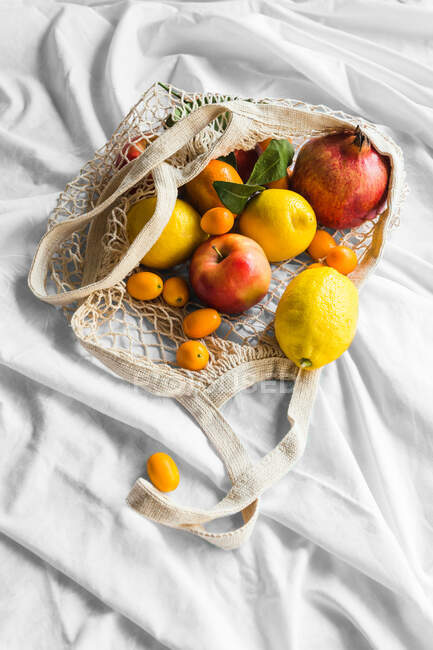 From above of different whole ripe apple with lemons near pomegranate and kumquats in eco bag on creased cloth - foto de stock
