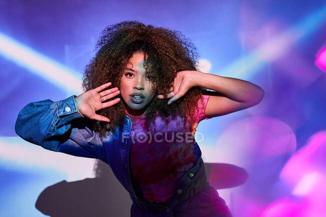 Attractive young African American female in casual wear touching curly hair and looking at camera while standing in dark studio in neon lights — Stock Photo