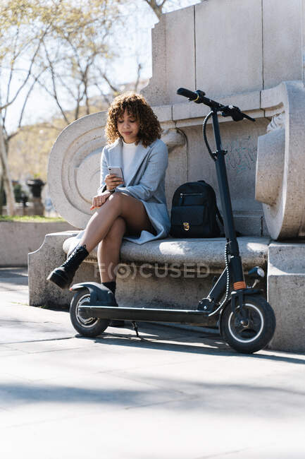 Full body young African American female in blue coat browsing on mobile phone while sitting on stone bench in spring park near scooter — Stock Photo