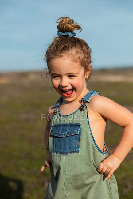 Очаровательная маленькая девочка в комбинезоне, стоящая с руками на талии на лугу и смотрящая вниз — стоковое фото