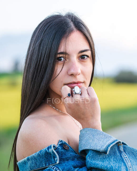 Gelassene junge Frau in abgesenkter Jeansjacke steht auf schmaler Fahrbahn in friedlicher Sommerlandschaft und blickt in die Kamera — Stockfoto