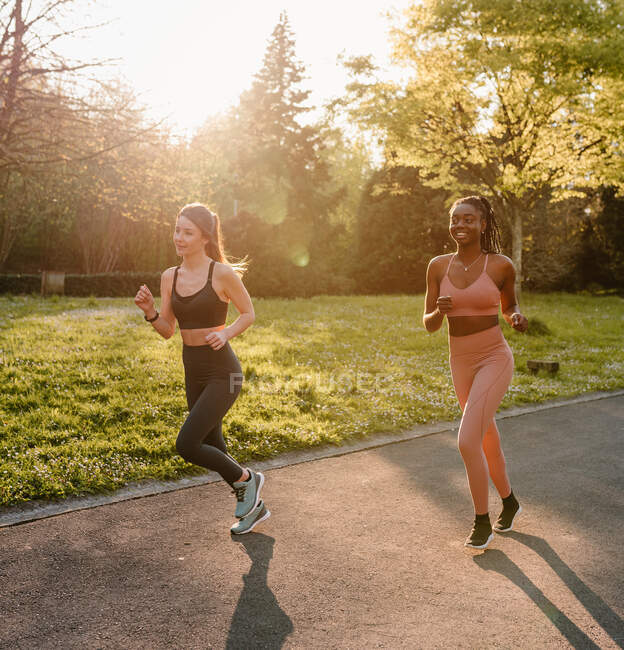 Sorridente corridori femminili multirazziali in activewear jogging e parlare durante l'allenamento cardio sulla passerella in città — Foto stock