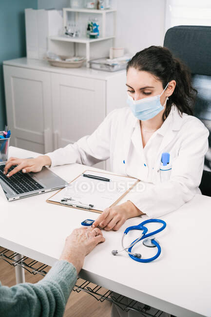 Young female doctor with laptop checking up heartbeat of crop anonymous patient with heart rate monitor at desk in hospital during pandemic — Stock Photo