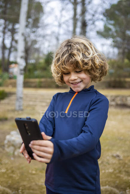 Cheerful child in blue wear with wavy hair taking self portrait on cellphone in daytime — Fotografia de Stock