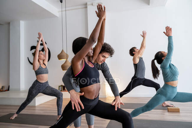 Vista laterale della compagnia di diverse persone che praticano yoga in posa Warrior con l'aiuto di allenatore in studio — Foto stock
