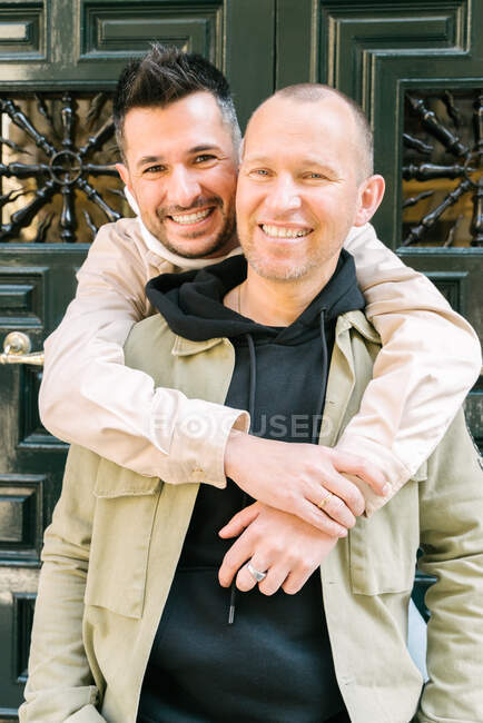 Fröhliche junge homosexuelle Männer in stilvollen Outfits, die lächelnd und umarmend auf der Straße vor der Tür stehen und in die Kamera schauen — Stockfoto