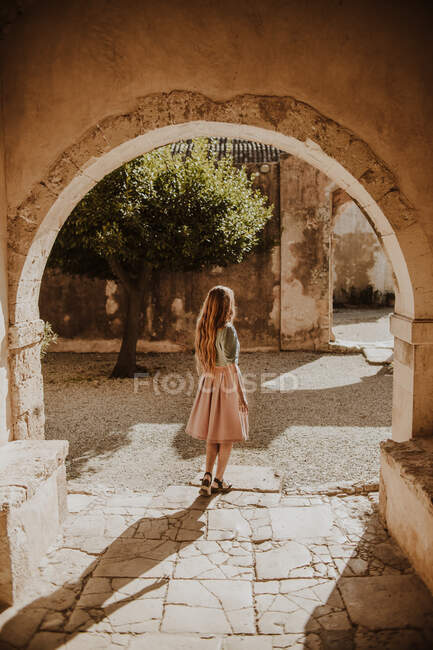Vista trasera hembra irreconocible en vestido de verano con estilo de pie cerca del arco del edificio medieval en el soleado día de verano en Heraklion Milos - foto de stock