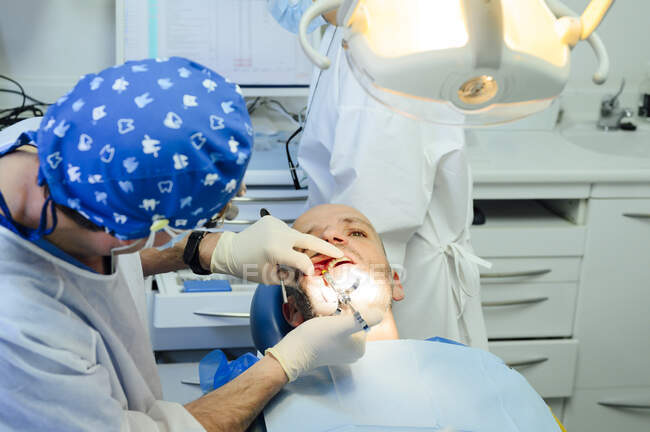 Seitenansicht eines anonymen Zahnarztes in Uniform und medizinischem Fernglas mit zufriedenem Mann auf Stuhl in der Klinik — Stockfoto