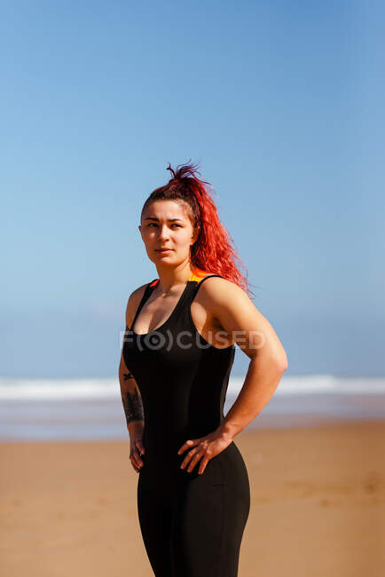 Самоуверенная спортсменка с руками на бедрах, смотрящая на песчаный пляж океана под солнечным светом — стоковое фото