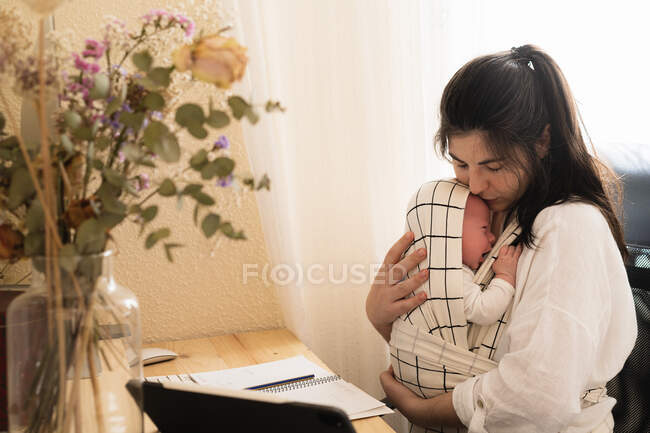 Mother embracing upset little child in baby carrier while sitting at table with flowers in vase at home — Stock Photo